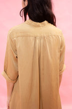 A woman viewed from behind wearing a tan linen shirt with rolled-up sleeves and a high mock neck, set against a solid pink background.