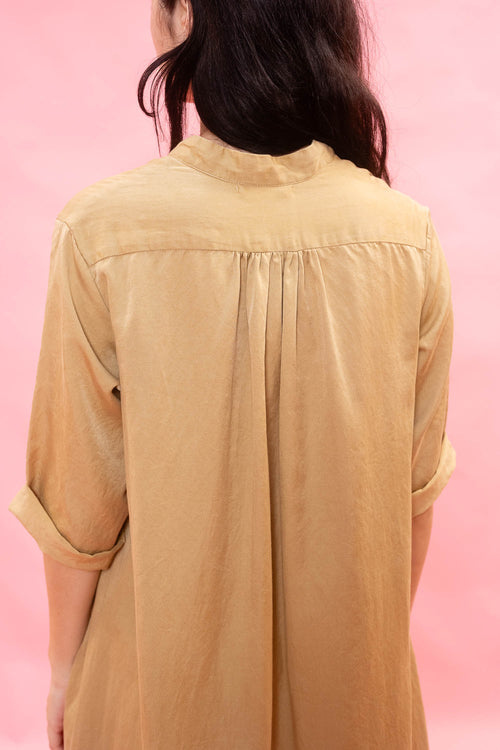 A woman viewed from behind wearing a tan linen shirt with rolled-up sleeves and a high mock neck, set against a solid pink background.