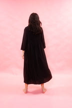 A woman viewed from behind wearing a long black midi dress with three-quarter sleeves, standing barefoot against a solid pink studio backdrop.