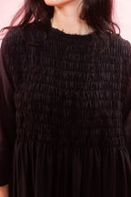 A close-up of a woman wearing a black smocked three-quarter sleeve dress against a pink background.