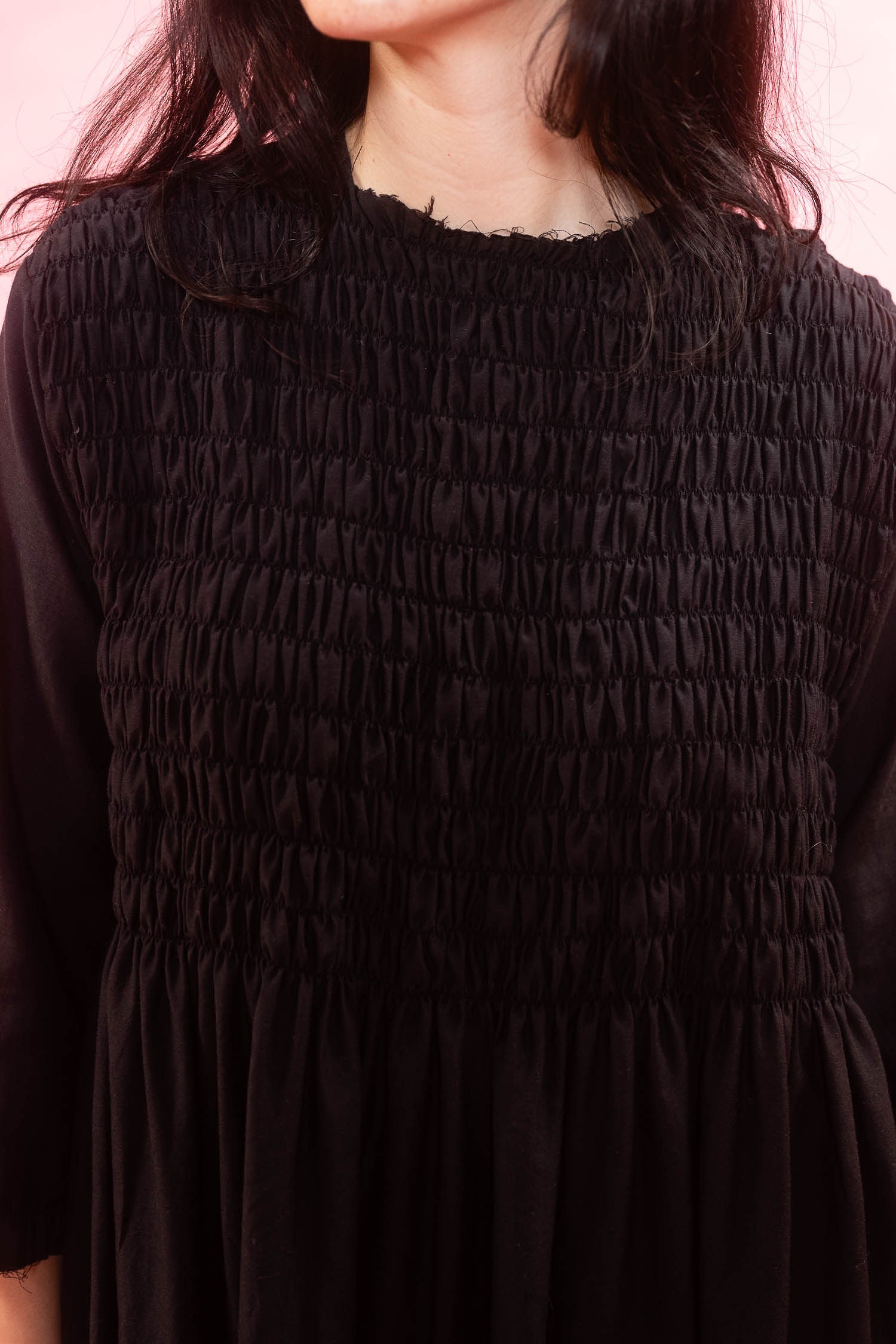 A close-up of a woman wearing a black smocked three-quarter sleeve dress against a pink background.