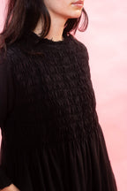 A close-up of a woman wearing a black smocked dress against a soft pink background.