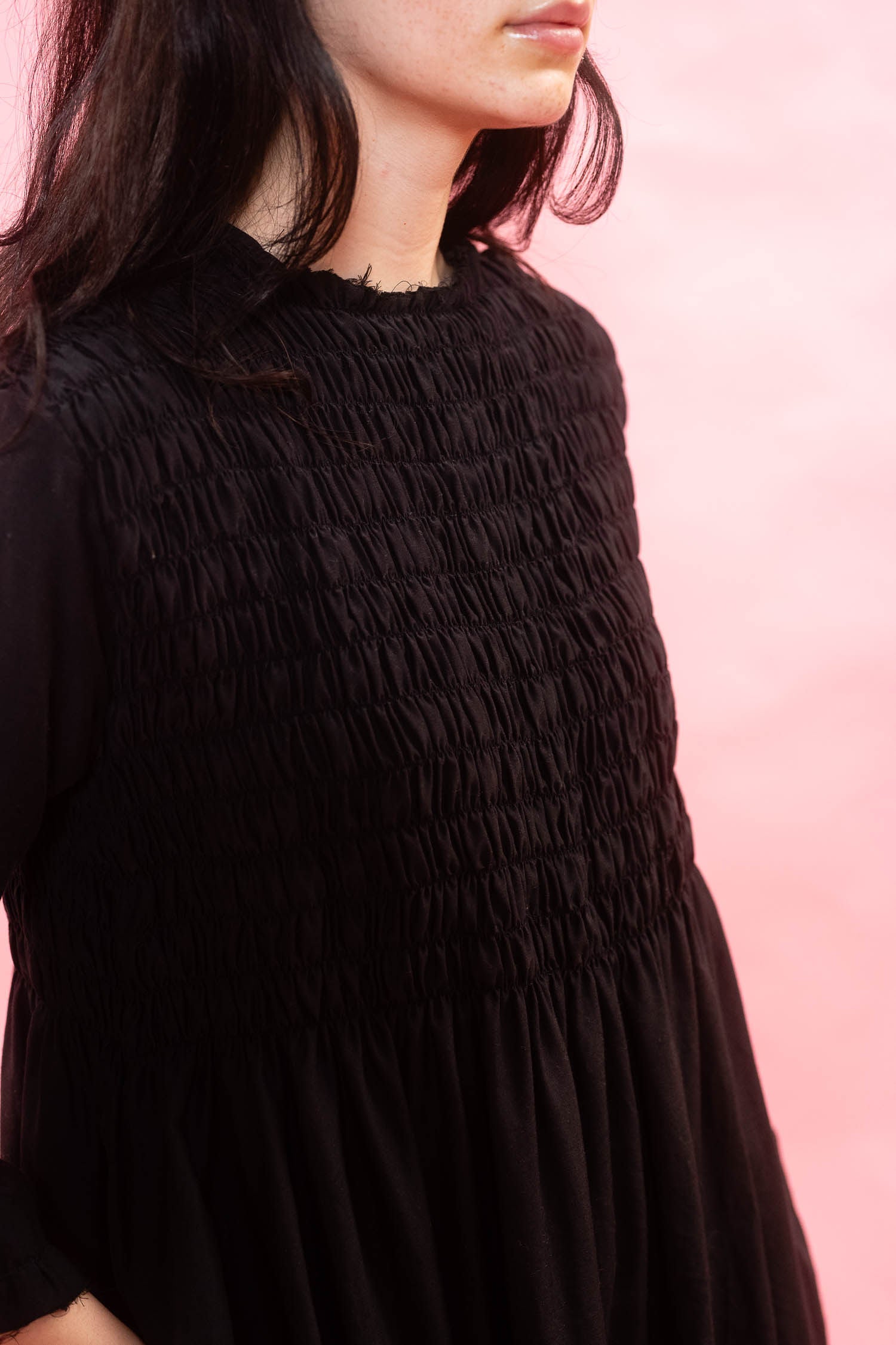 A close-up of a woman wearing a black smocked dress against a soft pink background.