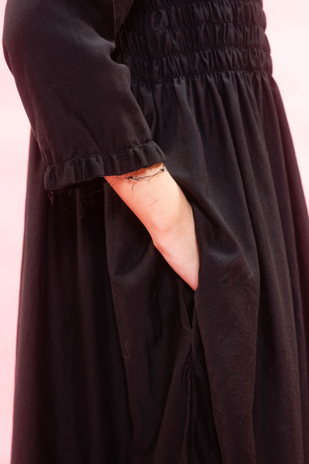 Close-up of a black smocked dress with a ruffled three-quarter sleeve and side pockets, set against a solid pink background.