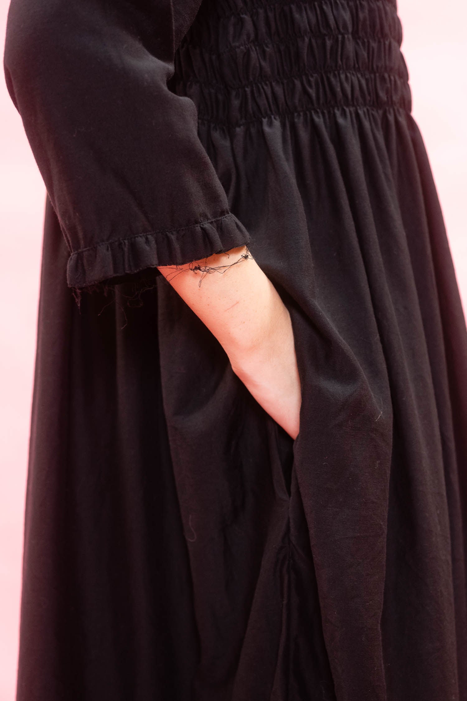 Close-up of a black smocked dress with a ruffled three-quarter sleeve and side pockets, set against a solid pink background.