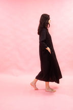 A woman wearing a long black dress with side pockets walks barefoot in profile against a solid pink studio backdrop.