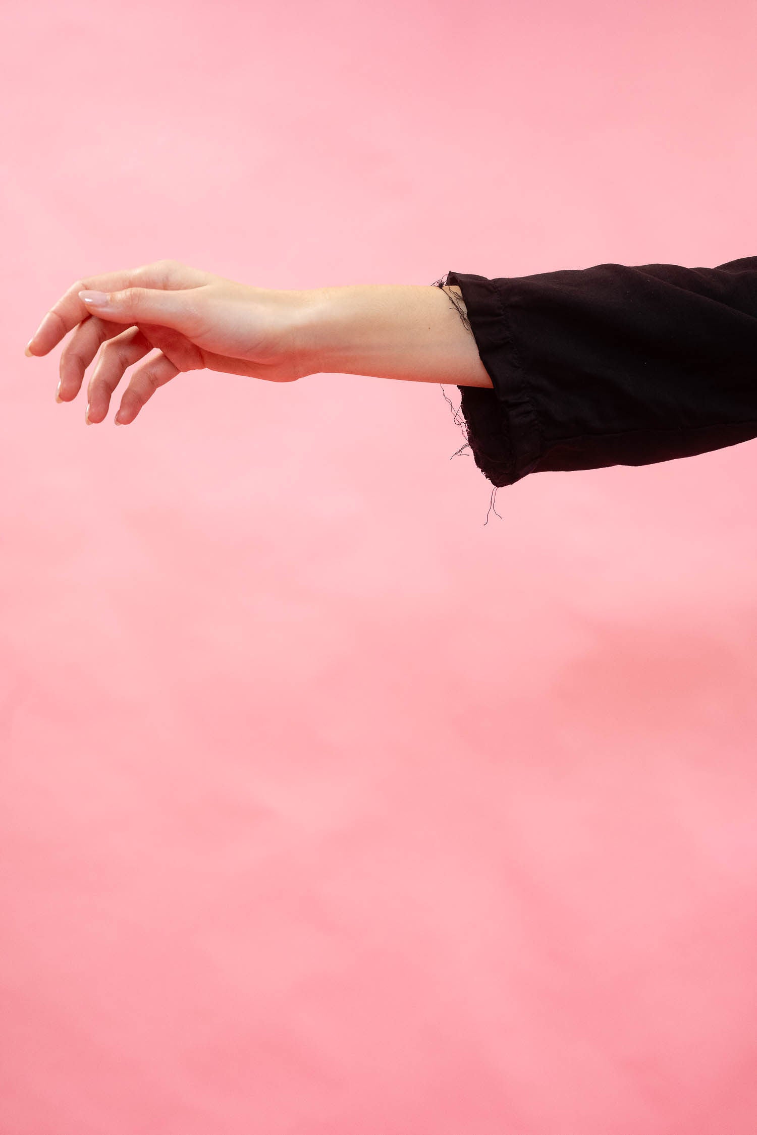 A woman's outstretched hand and forearm wearing a raw edged black sleeved dress, set against a solid pink background.