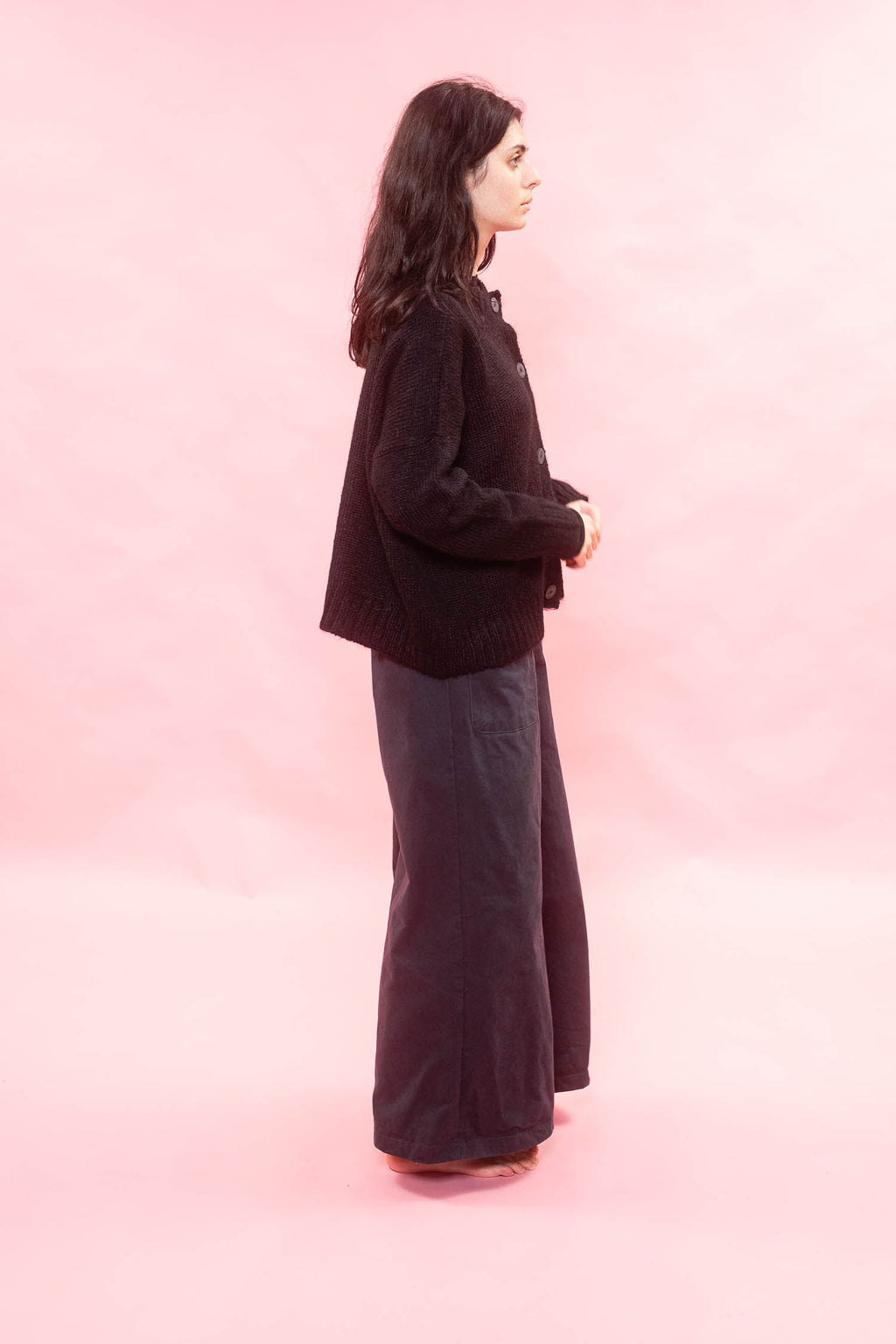 A woman stands in profile wearing a black  knit alpaca cardigan and dark wide-leg trousers against a solid pink studio background.