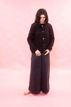 A woman stands barefoot against a solid pink studio background, wearing a black button-front alpaca 
cardigan and matching dark wide-leg pants.