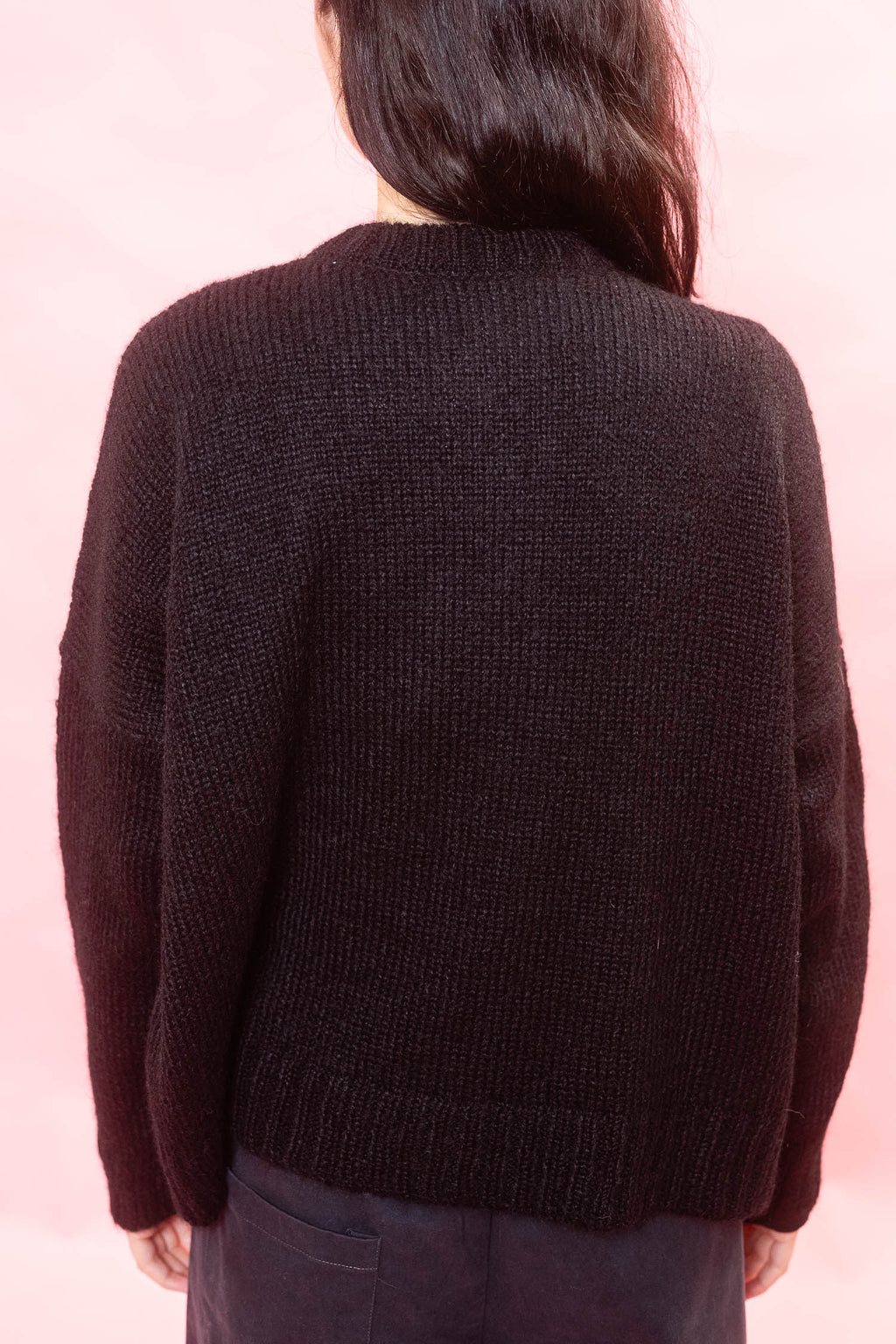 A person viewed from the back wearing a black alpaca cardigan and dark pants against a soft pink background.