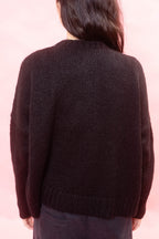 A person viewed from the back wearing a black alpaca cardigan and dark pants against a soft pink background.