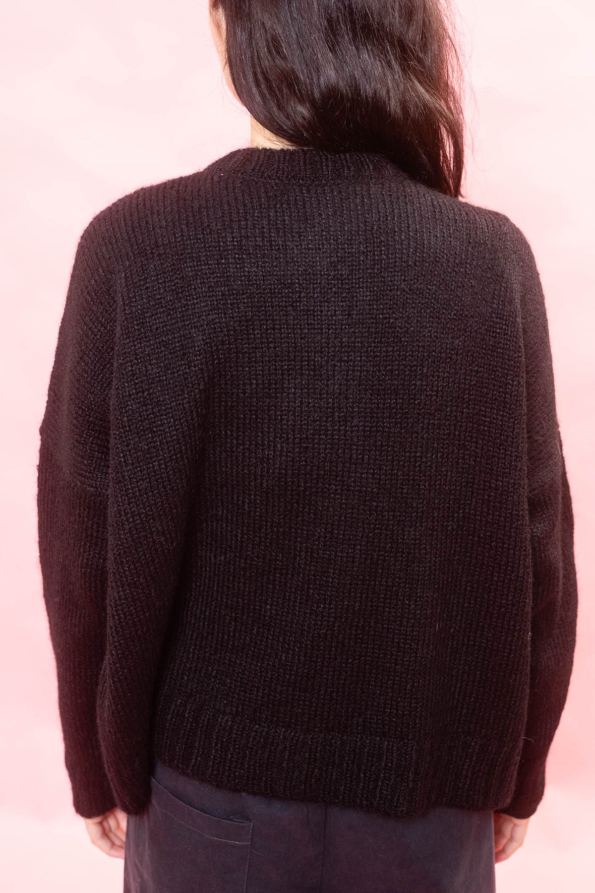 A person viewed from the back wearing a black alpaca cardigan and dark pants against a soft pink background.