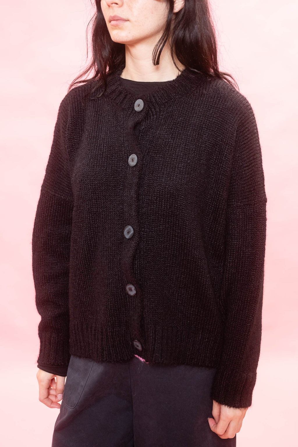 A person wearing black fluffy knit alpaca cardigan with dark wood buttons, paired with dark pants, set against a solid pink background.