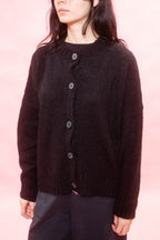 A person wearing black fluffy knit alpaca cardigan with dark wood buttons, paired with dark pants, set against a solid pink background.