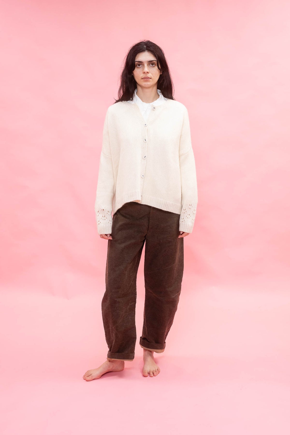 A woman stands barefoot against a solid pink studio backdrop, wearing a cream button-front cashmere hand knit oversized cardigan over a white collared shirt and dark brown corduroy pants.
