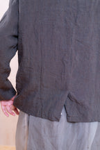 Close-up of a person wearing a dark charcoal gray linen long-sleeve top and light gray pants, showing the back of the shirt with a hand adjusting it.