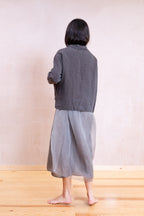 A person shown from the back wearing a dark gray cropped jacket and a loose light gray skirt, standing barefoot on a light wood floor against a plain beige wall.