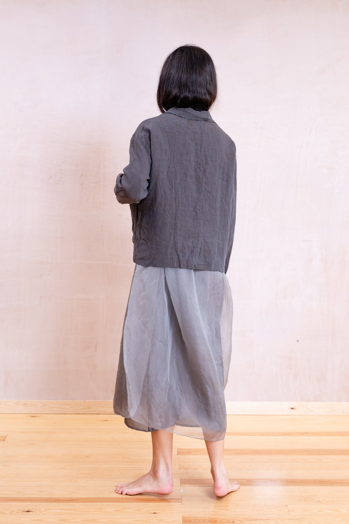 A person shown from the back wearing a dark gray cropped jacket and a loose light gray skirt, standing barefoot on a light wood floor against a plain beige wall.