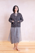A woman stands barefoot in a minimalist studio setting, wearing a dark charcoal gray button-up jacket with front patch pockets over a gray long-sleeve top and a matching sheer gray midi skirt.