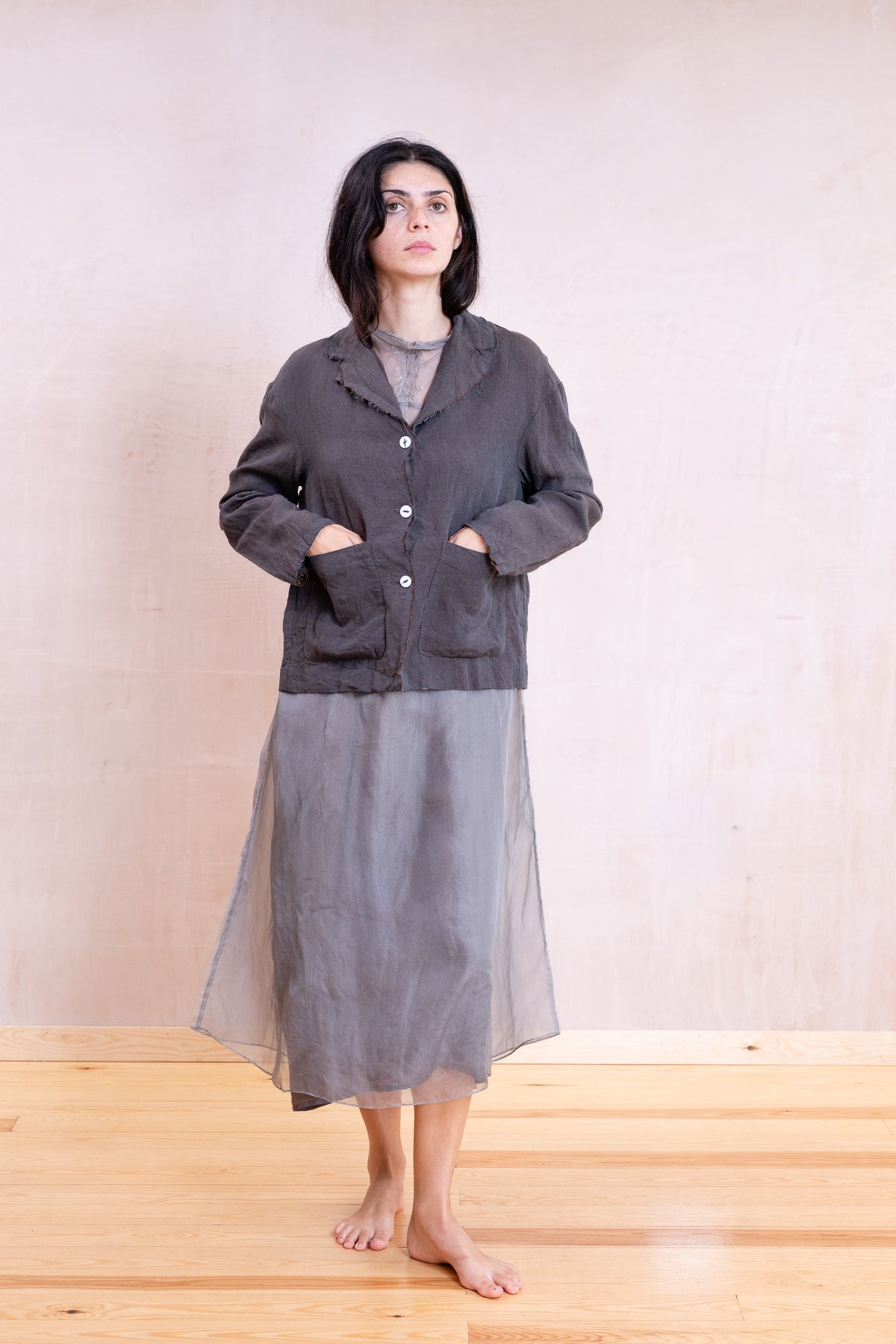 A woman stands barefoot in a minimalist studio setting, wearing a dark charcoal gray button-up jacket with front patch pockets over a gray long-sleeve top and a matching sheer gray midi skirt.