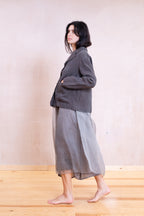 A woman stands barefoot in profile wearing a dark gray linen jacket and a sheer gray midi skirt against a light beige wall and wooden floor.