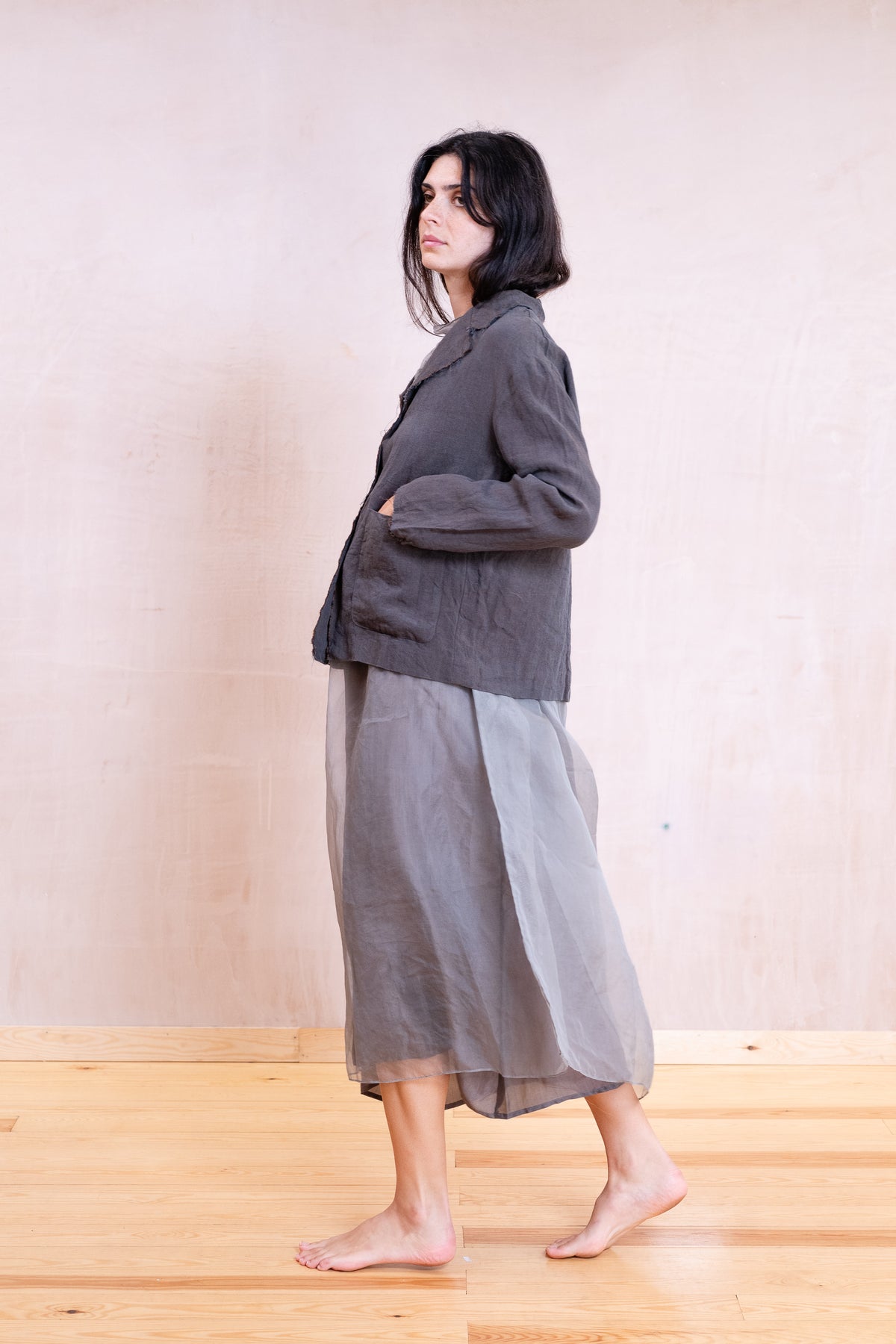 A woman stands barefoot in profile wearing a dark gray linen jacket and a sheer gray midi skirt against a light beige wall and wooden floor.