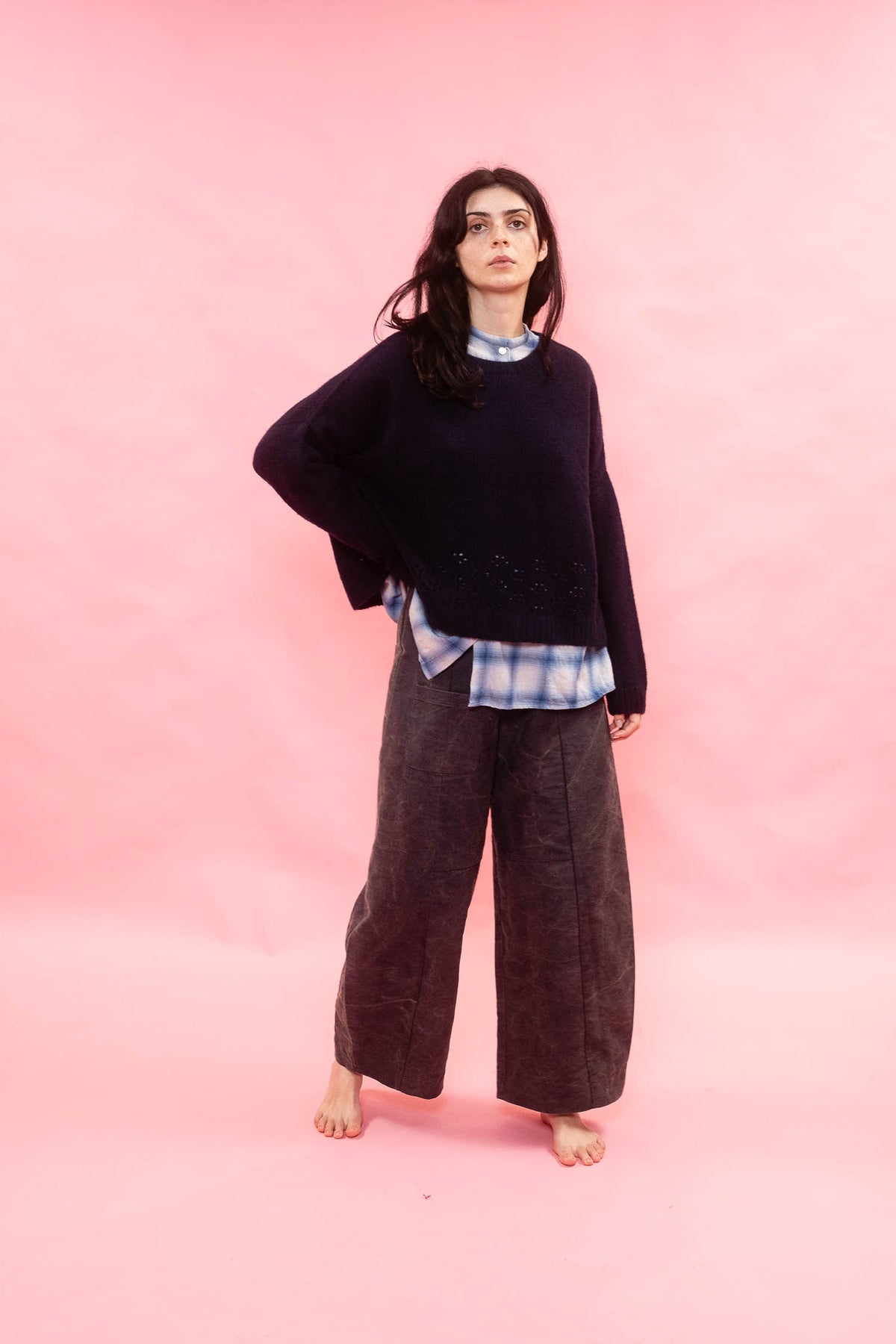 A woman stands barefoot against a solid pink studio backdrop, wearing a dark oversized knit sweater over a blue-and-white plaid shirt, paired with dark wide-leg trousers.