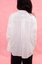 A person viewed from the back wearing a loose white long-sleeve button-up shirt and dark pants, standing against a solid pink backdrop.