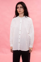 A woman with dark hair wearing a white long-sleeve button-up shirt and black pants, standing against a solid pink background.