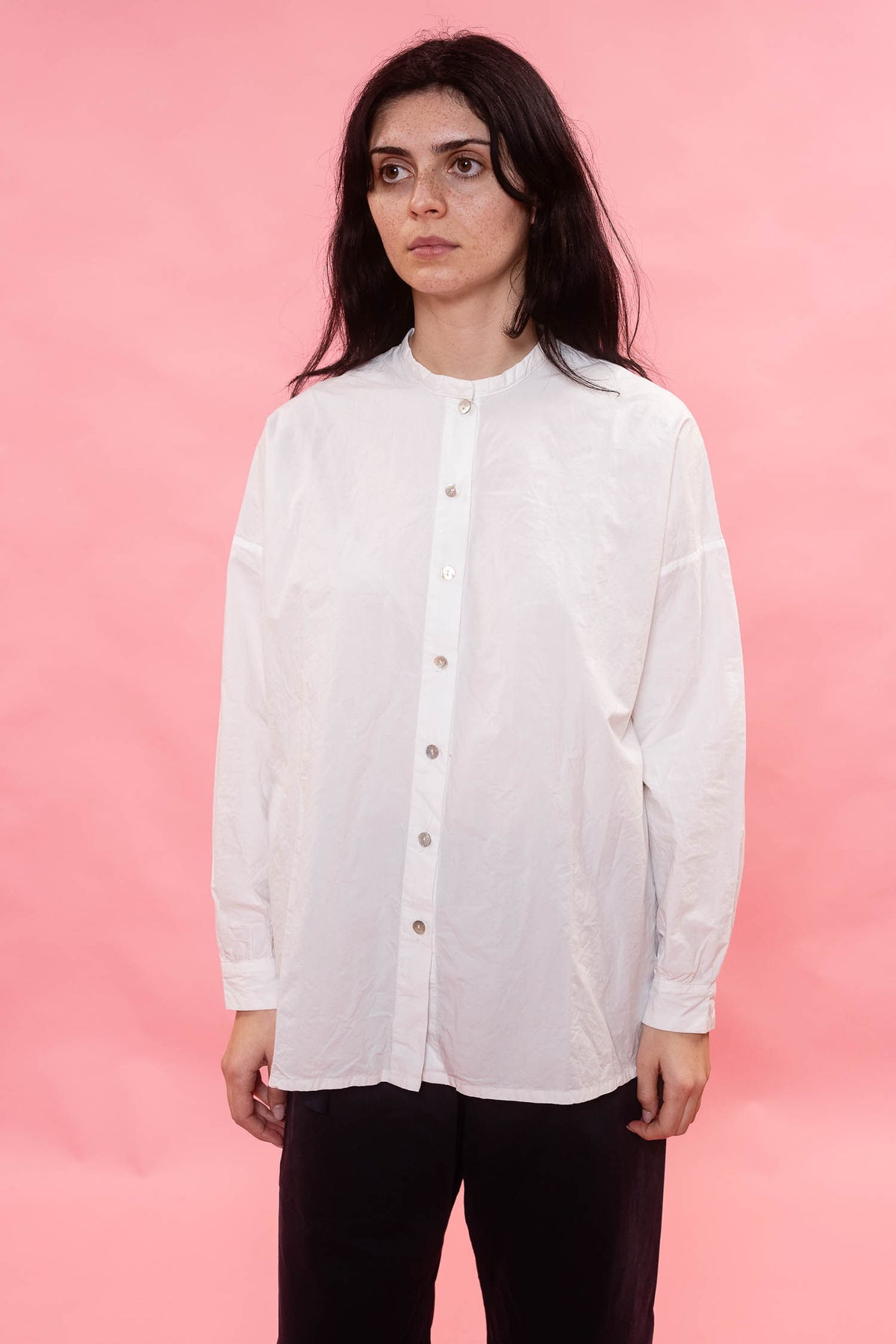 A woman with dark hair wearing a white long-sleeve button-up shirt and black pants, standing against a solid pink background.