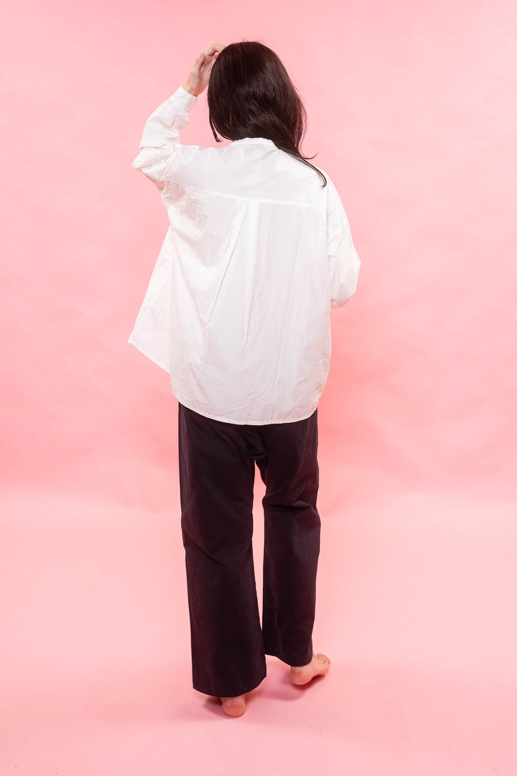 A person shown from behind wearing a loose white long-sleeve shirt and dark wide-leg pants, standing barefoot against a solid pink studio background.
