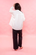 A person shown from behind wearing a loose white long-sleeve shirt and dark wide-leg pants, standing barefoot against a solid pink studio background.