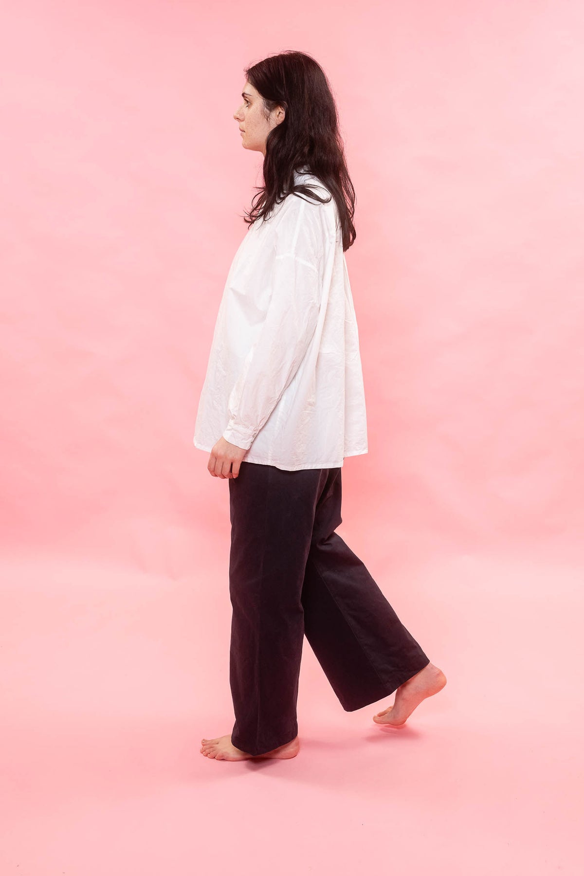 A woman walking in profile wearing a loose white long-sleeve shirt and dark wide-leg pants against a solid pink studio background.