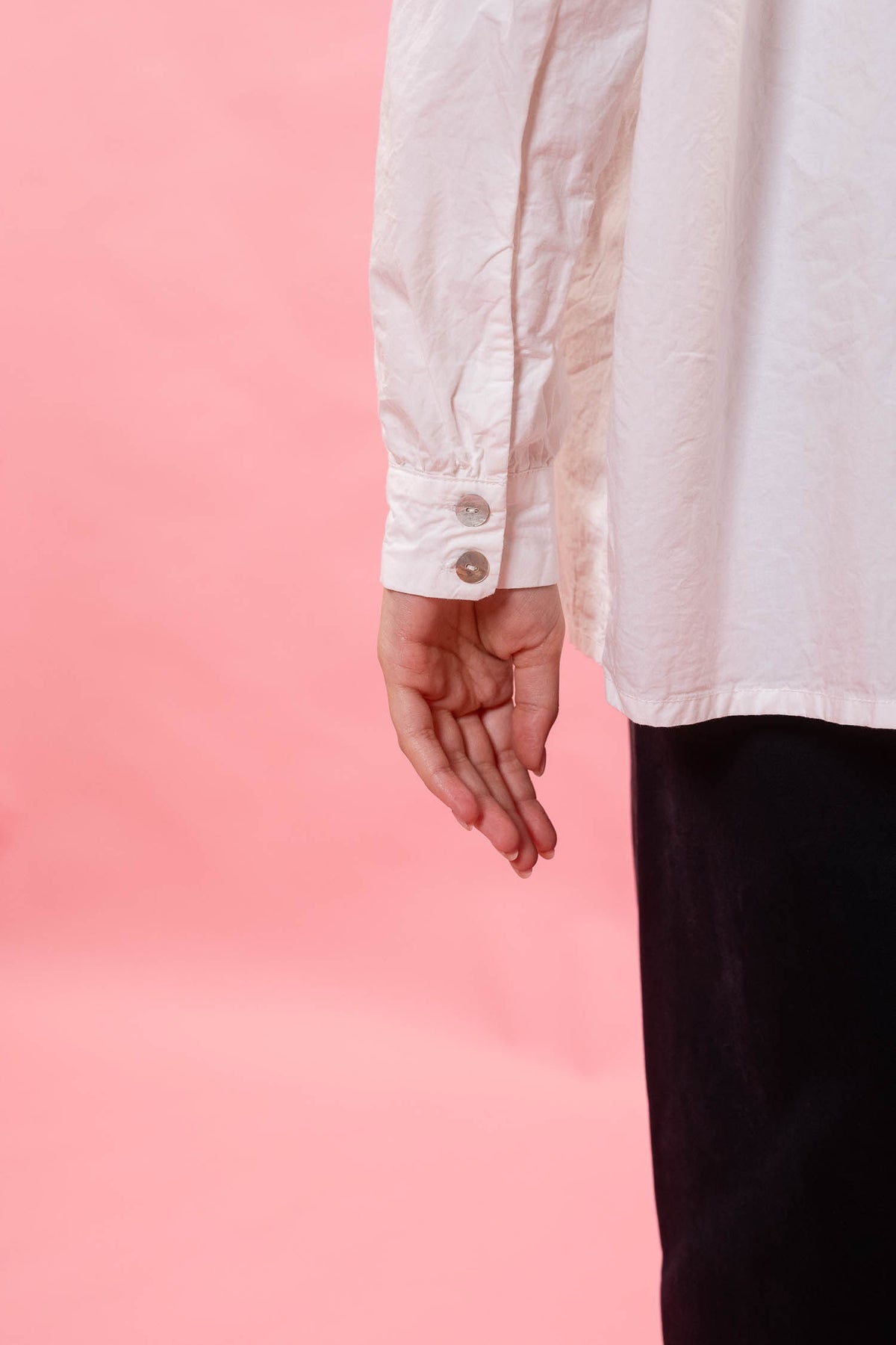 A close-up of a person wearing a white long-sleeve button-up shirt and dark pants, with the hand and lower torso visible against a solid pink background.