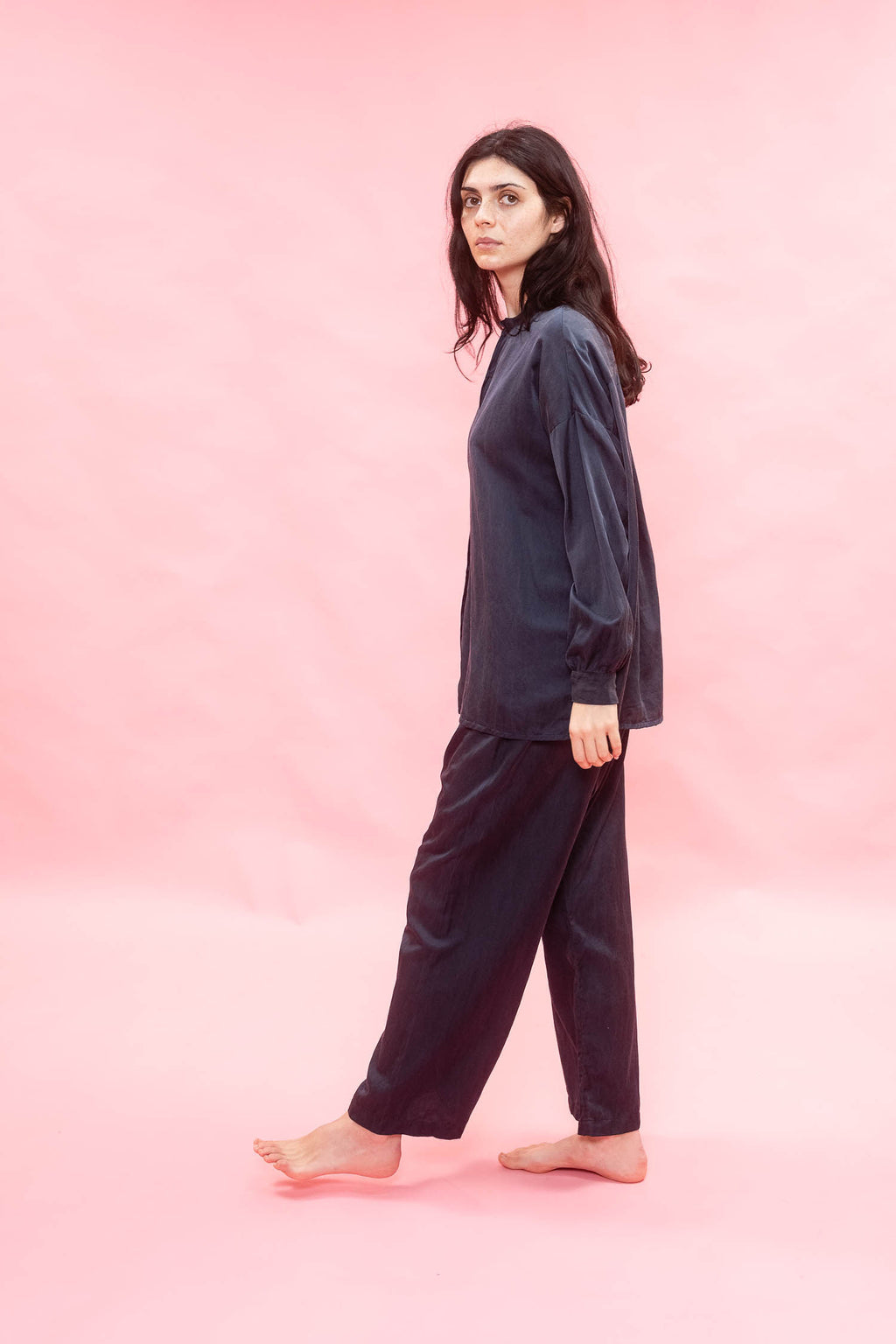 A woman modeling a dark navy blue two-piece pajama set consisting of a long-sleeve top and matching pants, standing against a solid pink studio backdrop.
