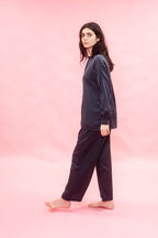A woman modeling a dark navy blue two-piece pajama set consisting of a long-sleeve top and matching pants, standing against a solid pink studio backdrop.