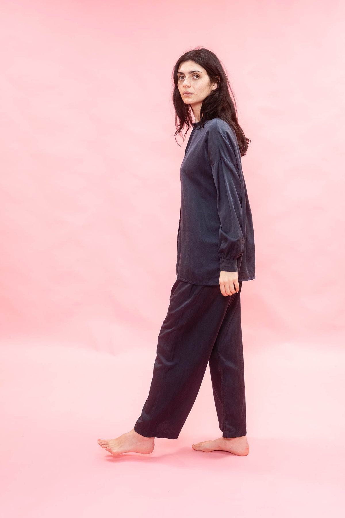 A woman modeling a dark navy blue two-piece pajama set consisting of a long-sleeve top and matching pants, standing against a solid pink studio backdrop.
