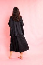 Back of model wearing a black silk shirt and black wool stripe skirt on a pink background