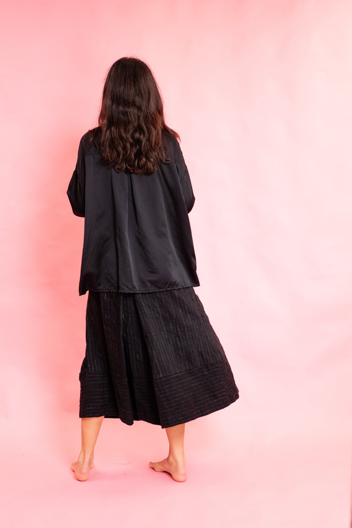 Back of model wearing a black silk shirt and black wool stripe skirt on a pink background
