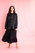 A woman stands barefoot against a solid pink studio backdrop, wearing a loose black long-sleeve button-front top and a black textured midi skirt.