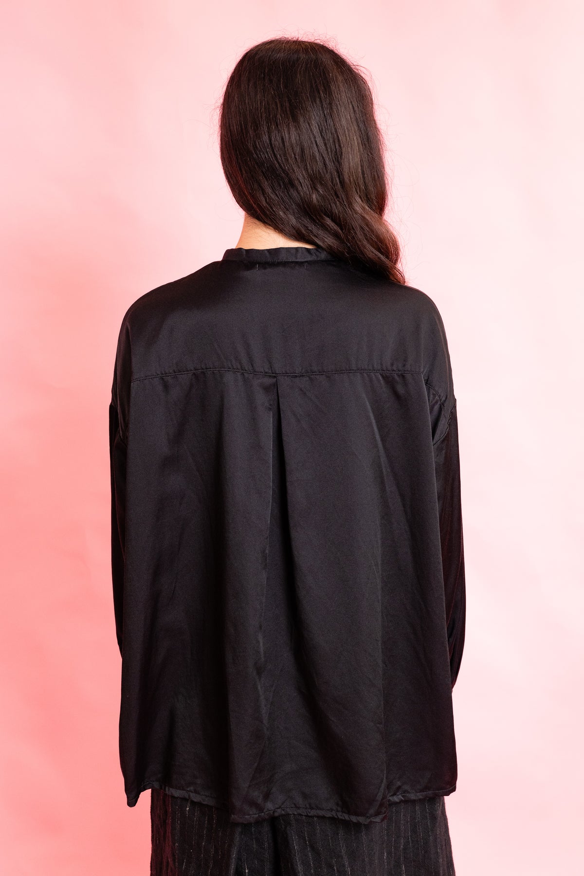 A woman viewed from behind wearing a loose black long-sleeve top and dark pleated bottoms, standing against a soft pink studio background.