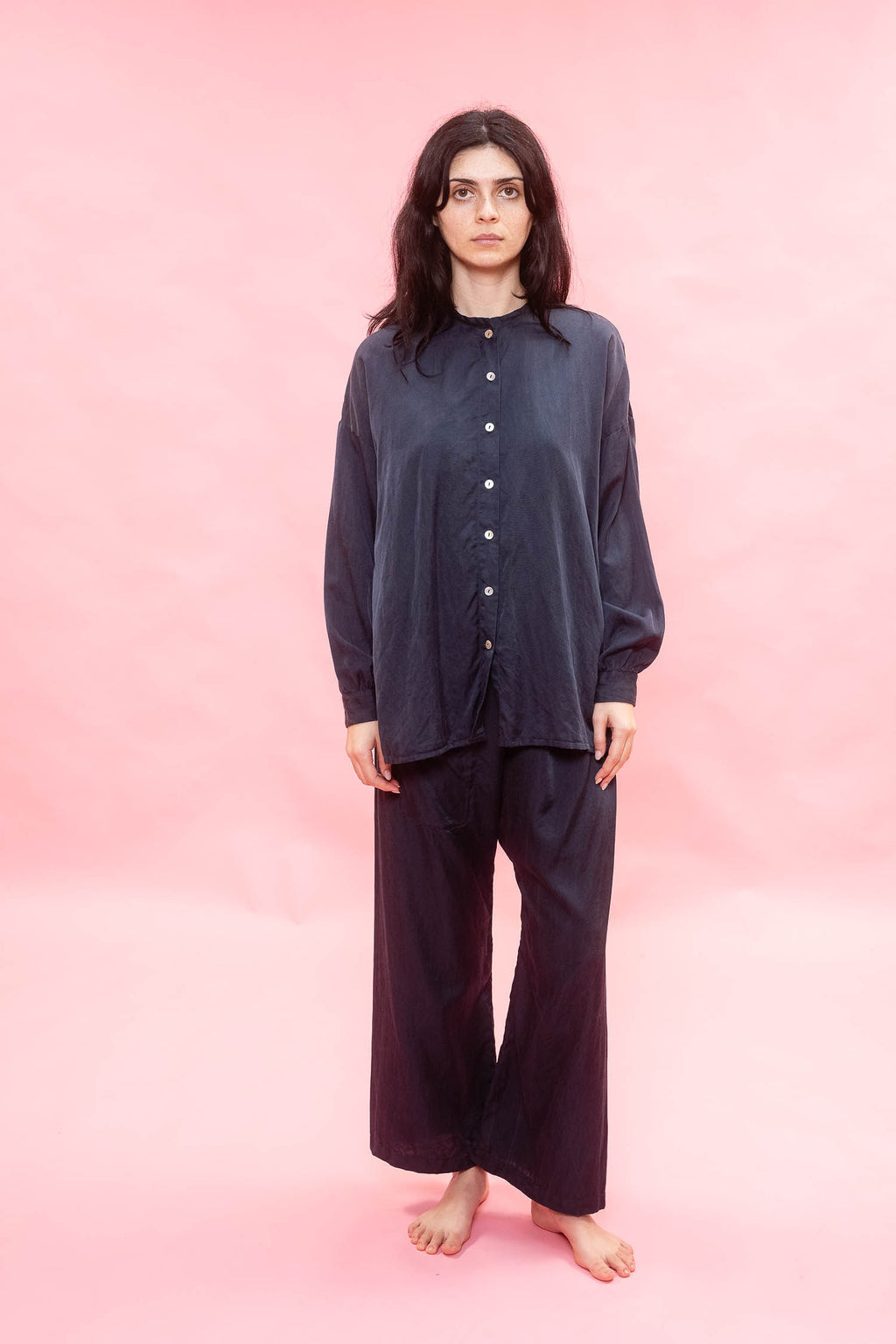 A woman stands barefoot against a solid pink studio backdrop, wearing a loose dark navy button-front top and matching wide-leg pants.