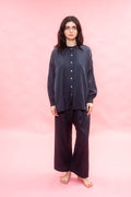 A woman stands barefoot against a solid pink studio backdrop, wearing a loose dark navy button-front top and matching wide-leg pants.