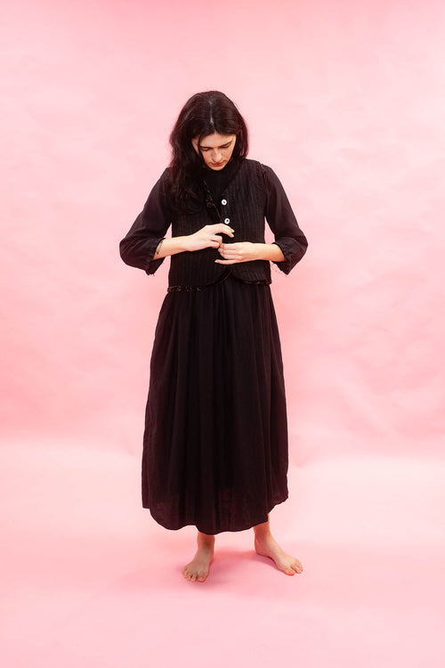 Woman wearing a black dress with a black softly striped wool linen waistcoat. standing against a pink background. 