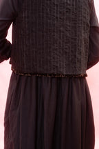 A close-up of a dark brown textured vest with vertical stripe details and a ruffled trim, worn over a matching dark brown long-sleeved dress against a light pink background.