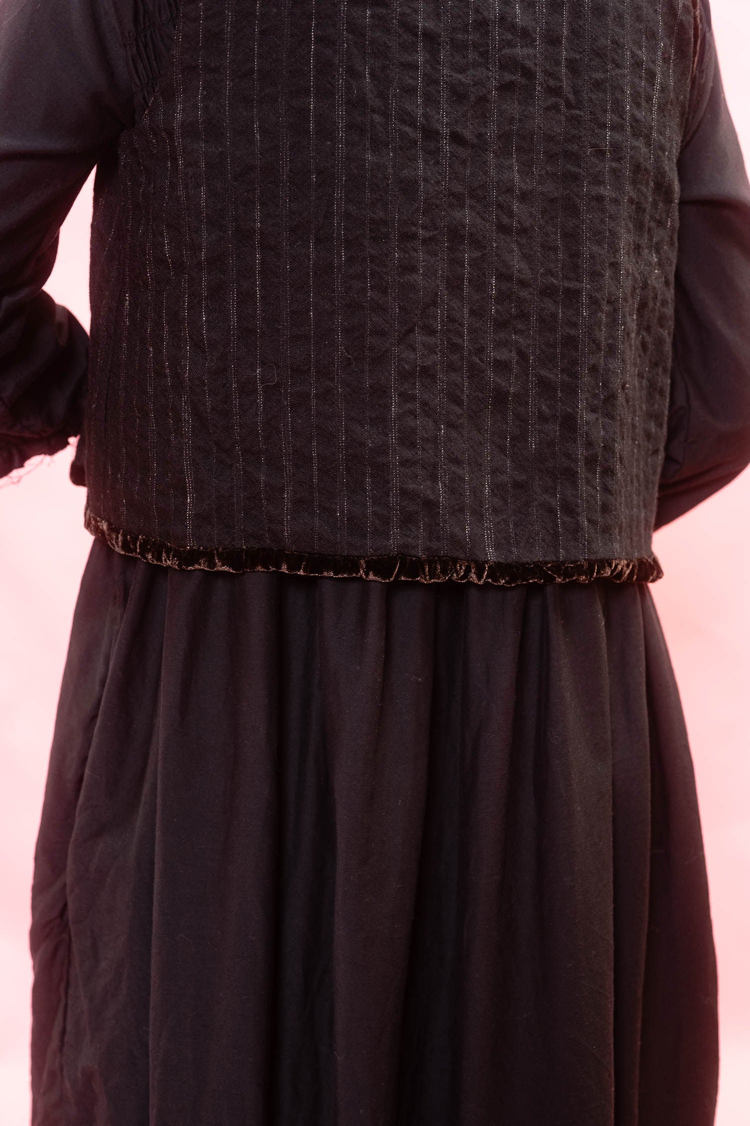 A close-up of a dark brown textured vest with vertical stripe details and a ruffled trim, worn over a matching dark brown long-sleeved dress against a light pink background.