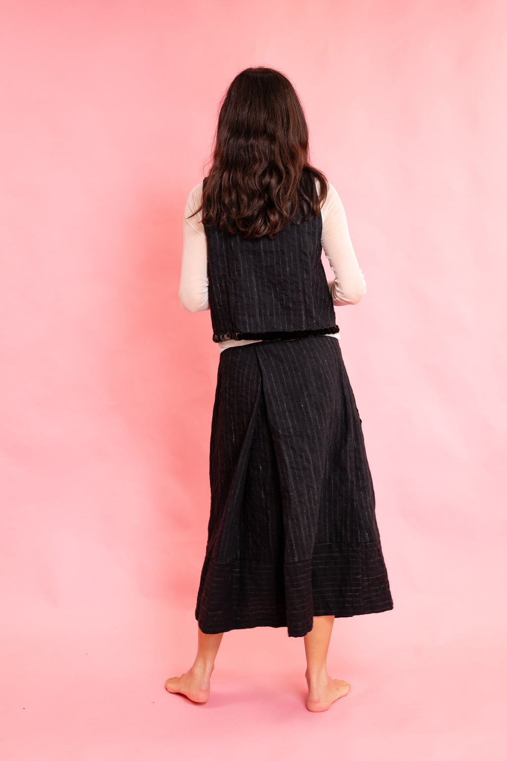 Back view of model wearing a wool stripe buttoned waistcoat with brown velvet trim with a cream mesh long sleeve tee shirt and panelled wool stripe skirt on a pink background