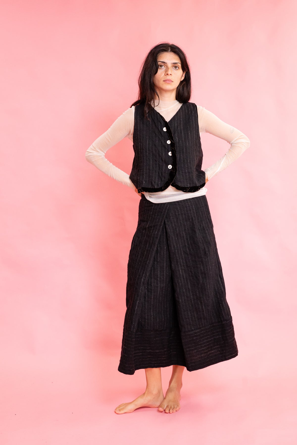 Model wearing a wool stripe buttoned waistcoat with brown velvet trim with a cream mesh long sleeve tee shirt and panelled wool stripe skirt on a pink background