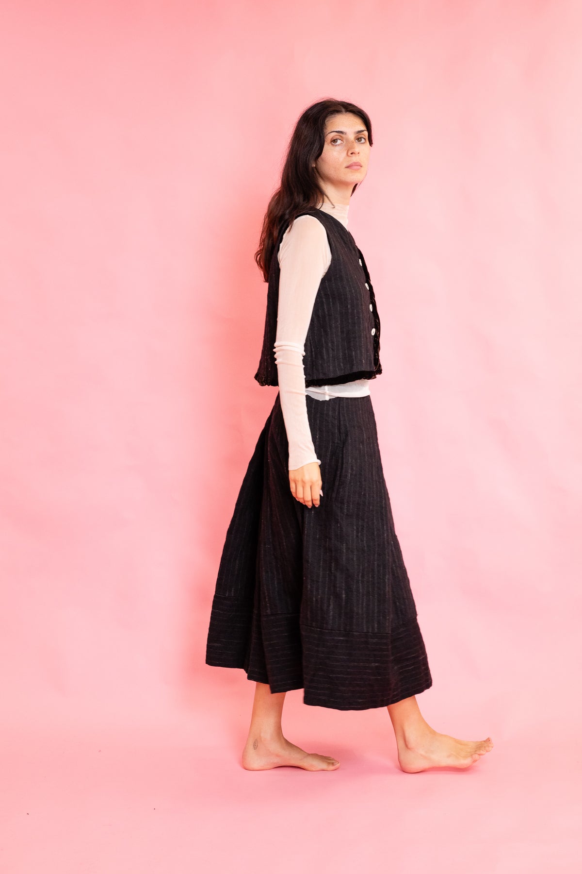 Side view of model wearing a wool stripe buttoned waistcoat with brown velvet trim with a cream mesh long sleeve tee shirt and panelled wool stripe skirt on a pink background