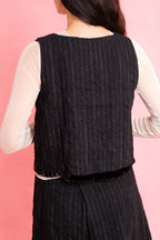 Back view of model wearing a wool stripe buttoned waistcoat with brown velvet trim with a cream mesh long sleeve tee shirt and panelled wool stripe skirt on a pink background